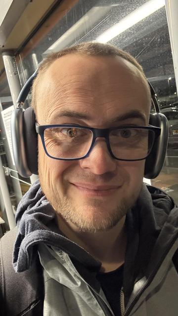 A smiling individual wearing headphones and glasses, with a short beard, is seen in a well-lit indoor setting with glass windows and a blurred background.